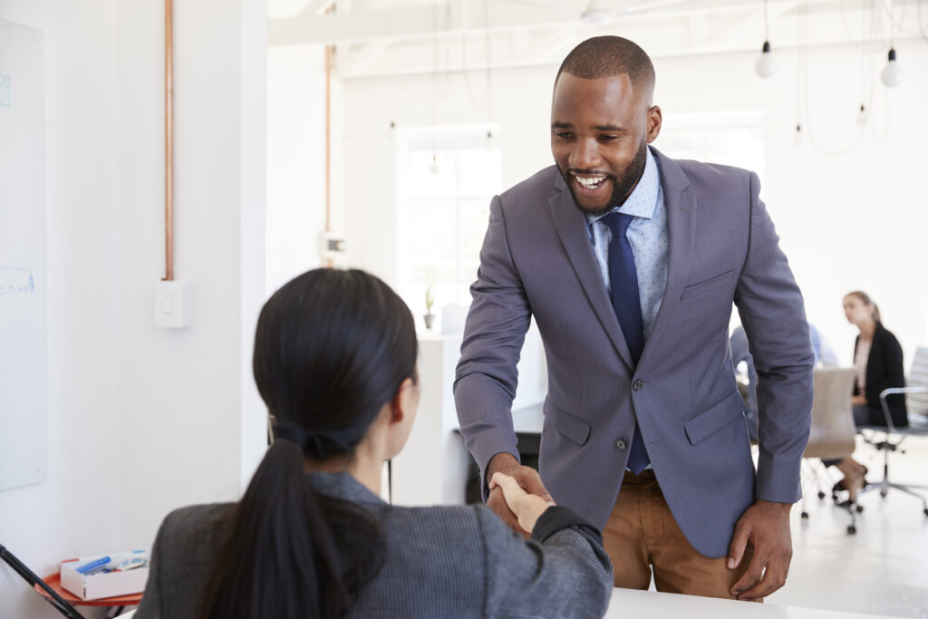 A young business professional shaking hands with his client, about to sit down for a meeting.