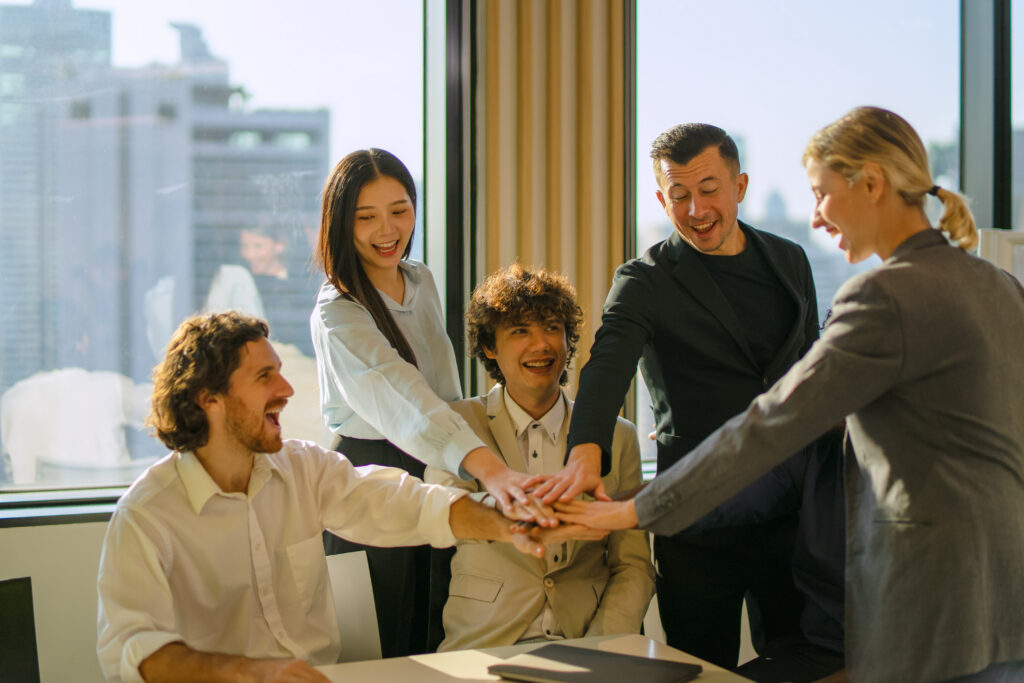 Five young professionals joining hands in a bright, indoor work environment.