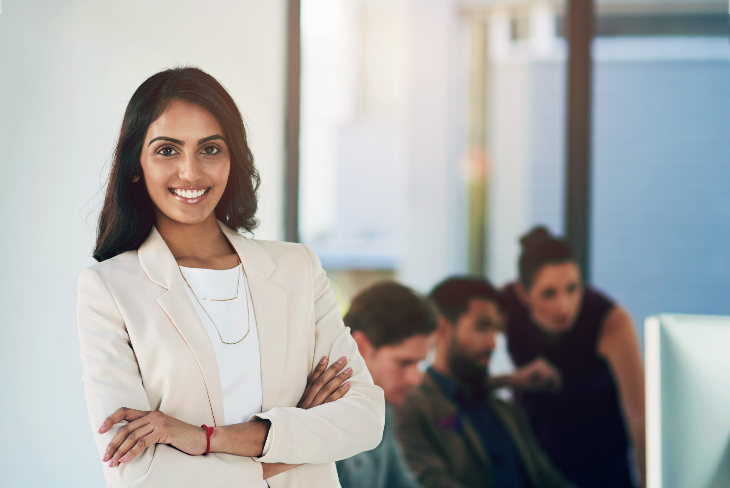 A smiling, confident young professional standing in a modern office setting with her colleagues working in the background.