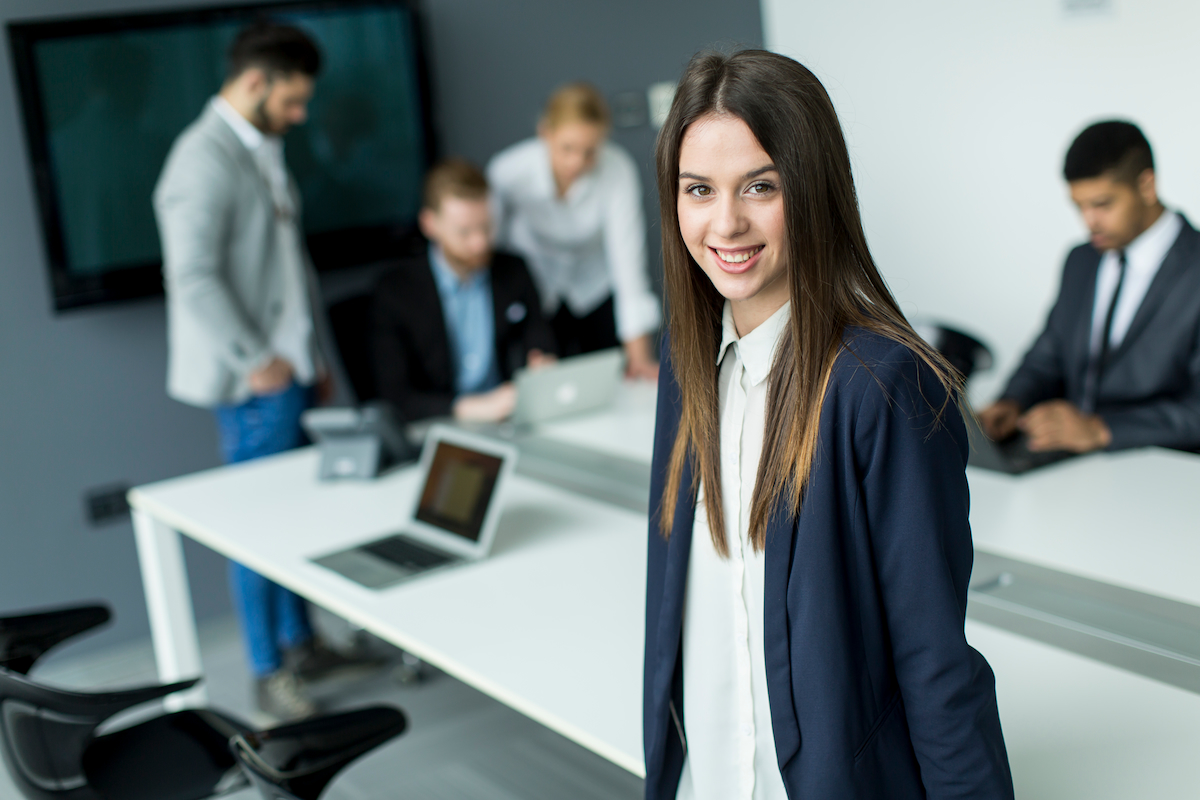 A young business professional in a modern office setting with her colleagues brainstorming behind her.